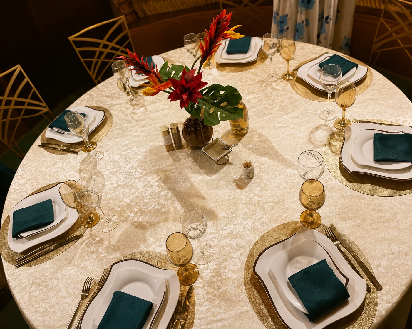 Table ronde dressée : sous-assiettes dorées, serviettes vert canard et fleurs tropicales